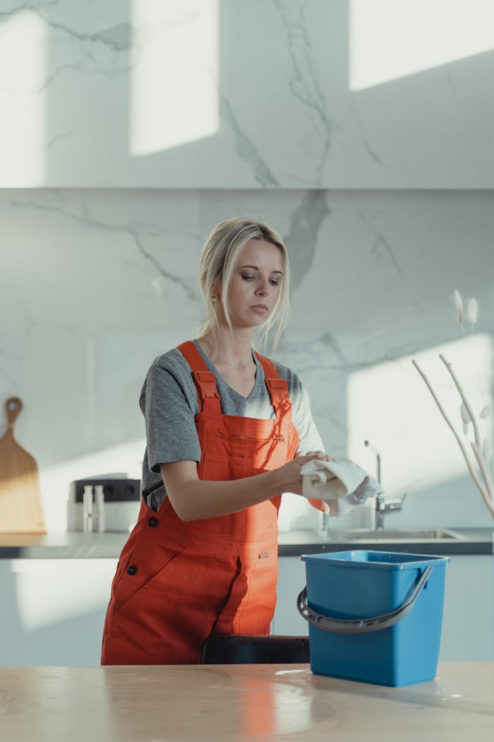 services-01 A woman wearing orange overalls cleans a kitchen counter with a rag and pail in a modern kitchen.